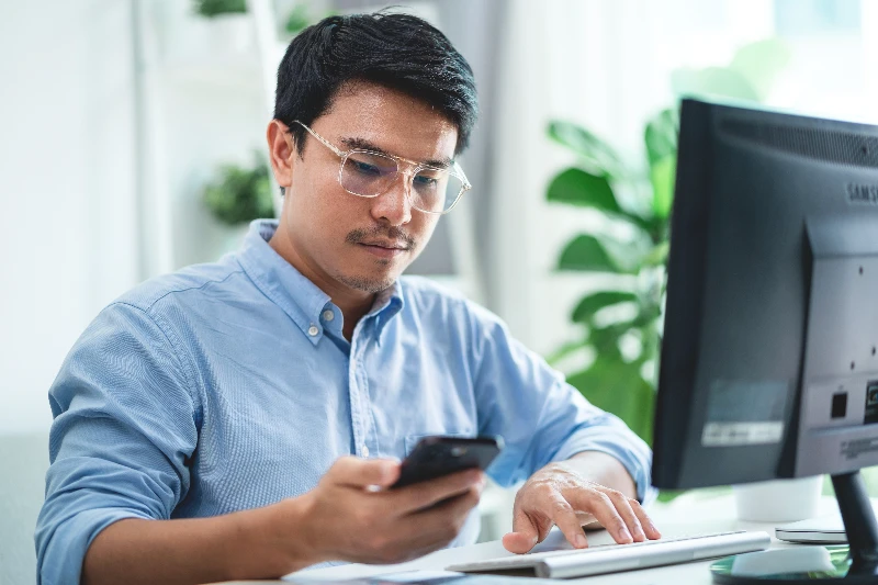 A man wearing glasses is sitting at a desk with a computer monitor and a phone